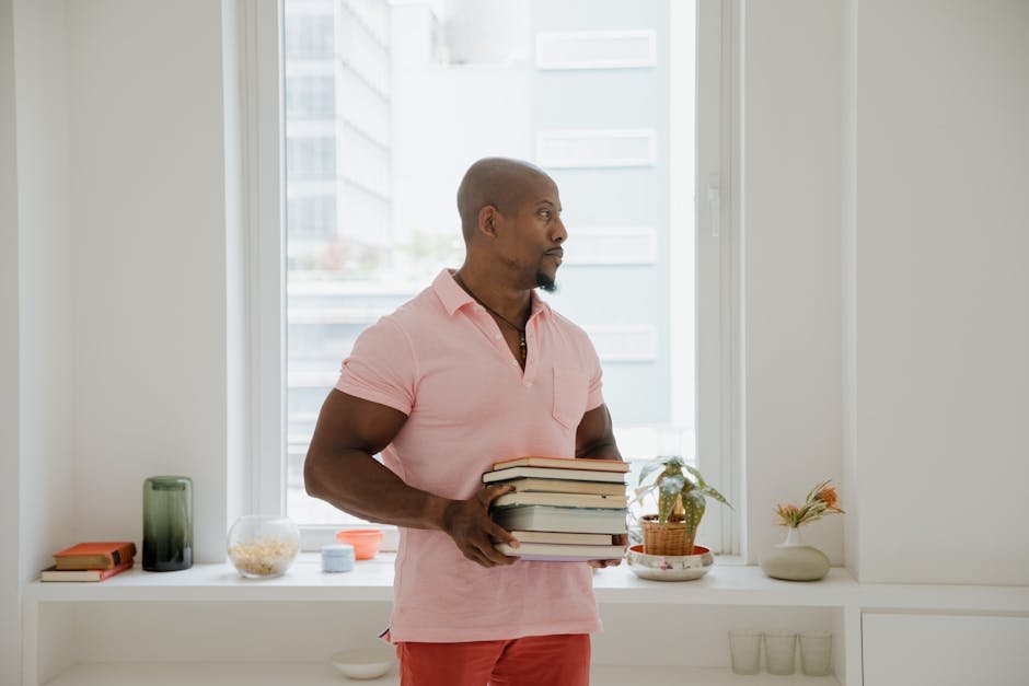 Man in pink shirt holding books indoors near window, conveying calm and focus.