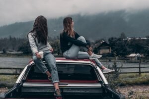 Two young women sitting on a car, enjoying the serene mountain landscape in Romania.