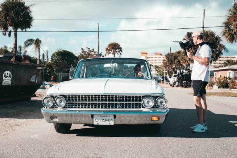 Filmmaker with handheld camera capturing a vintage car in a sunny urban street setting.