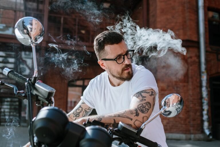 Stylish bearded man with tattoos riding a motorcycle, exhaling smoke outdoors.